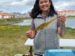 A woman stands near a seashore with a frond of kelp in her hands, and more kelp in containers on a table in front of her.
