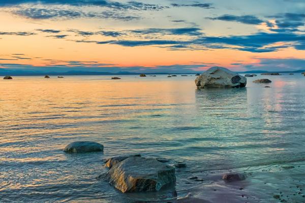 Blue and orange hued sky over distant snow capped mountains are in the background of large smooth rocks on an ocean beach with gentle waves.
