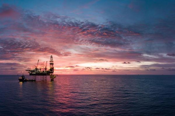 An ship anchored near an oil right at late sunset, both vessels have white lights turned on and are contrasted against a magnificent pink, purple, and blue sunset