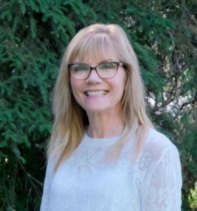 A woman with shoulder length blonde hair and glasses smiling at the camera, standing in front of evergreen trees.