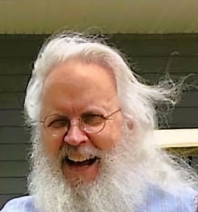 A man with glasses, long, white hair, and a long white beard grinning warmly towards a camera