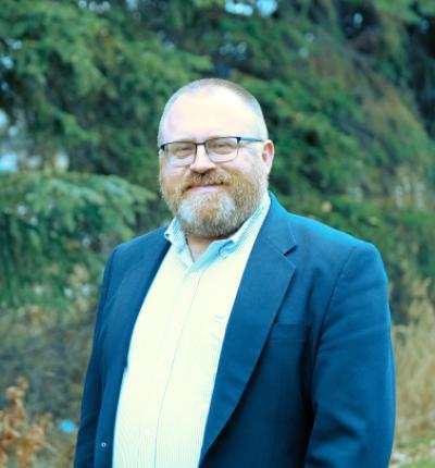 Portrait of Givey Kochanowski wearing office casual clothes in front of evergreen trees.
