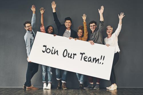 Studio shot of a group of people holding up a "join our team" placard against a grey background.