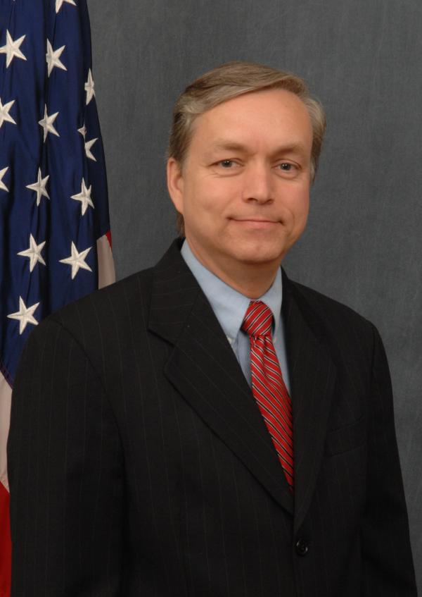 A man in a dark suit with a red tie standing just in front of a United States flag pole