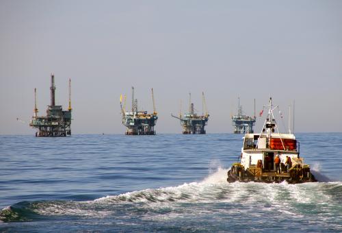 A boat is leaving a wake as it drives away from the camera towards four offshore rigs in the distance.