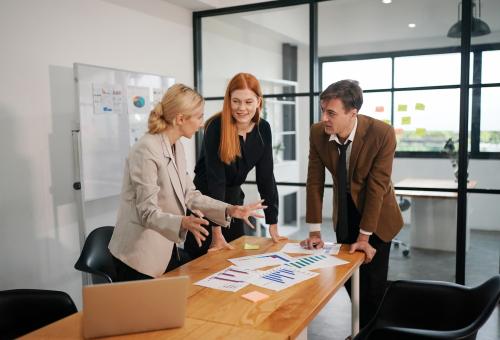 Three colleagues, two women and one man, review charts and graphs on a table in a modern office.